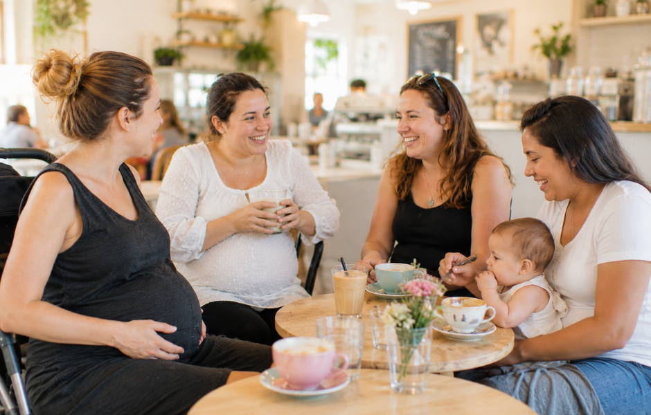 Imagen de cuatro mujeres, tres de ellas embarazadas y una con un bebé, sentadas alrededor de una mesa, tomando un café. Están dentro de una cafetería con mucha luz natural y un mobiliario y decoración muy cuidada
