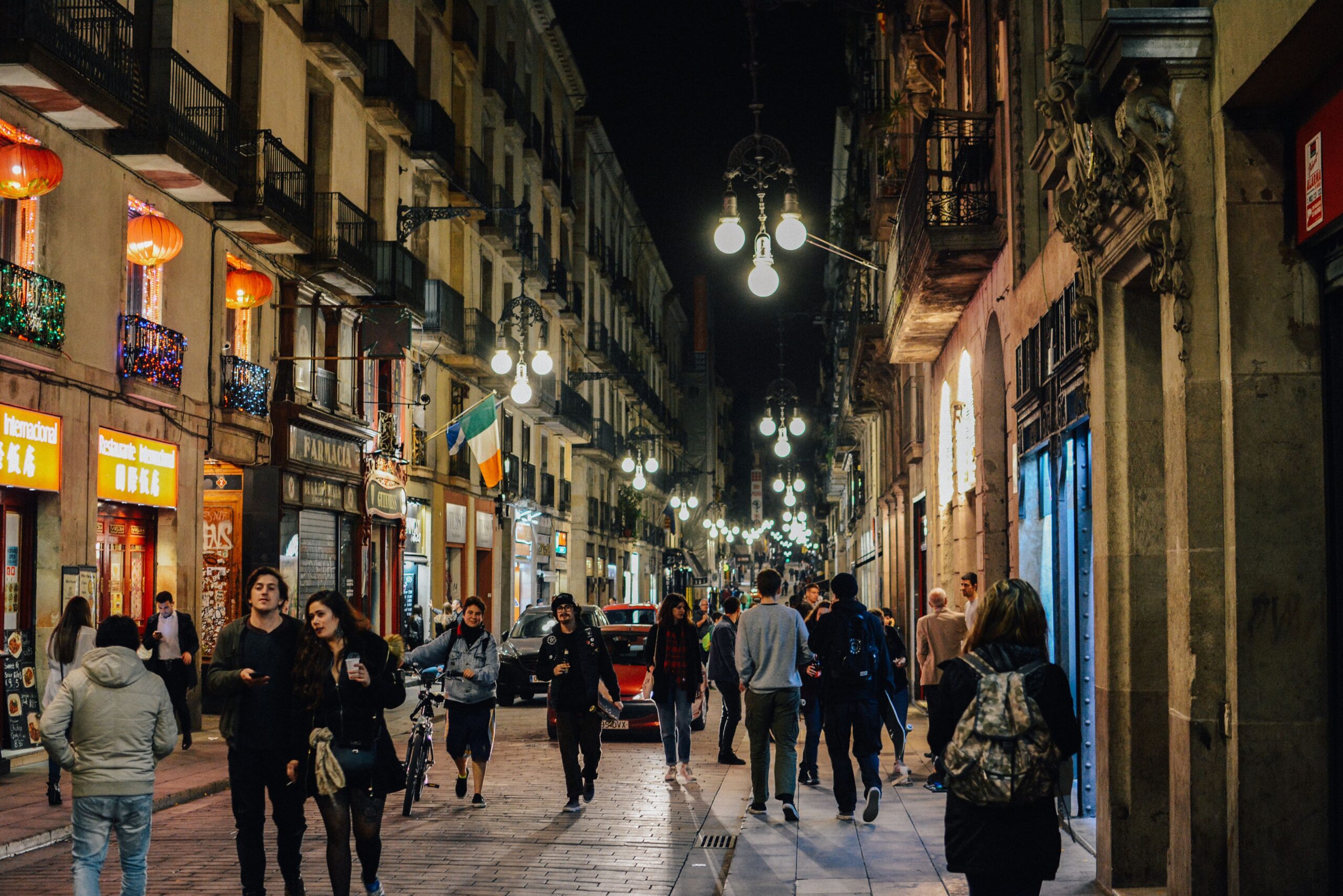 Una calle peatonal de ciudad al anochecer, iluminada por farolas, con gente caminando en ambos sentidos entre edificios antiguos con balcones y comercios.
